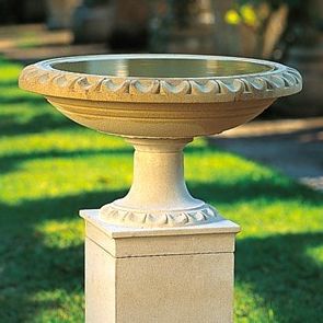 Ein großer, beiger Steinpflanzenbehälter mit dekorativen Kanten steht auf einem Betonweg inmitten eines Rasenbereichs.