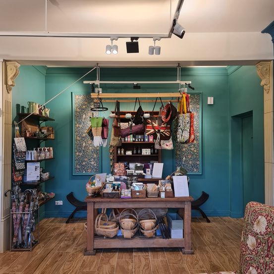 Innenansicht eines Ladens mit türkisfarbenen Wänden, Holzboden und verschiedenen ausgestellten Artikeln. An beiden Seiten befinden sich Regale mit Taschen, Büchern und Flaschen. Oben befindet sich eine Decke mit Strahlern und einem offenen Deckenbereich.