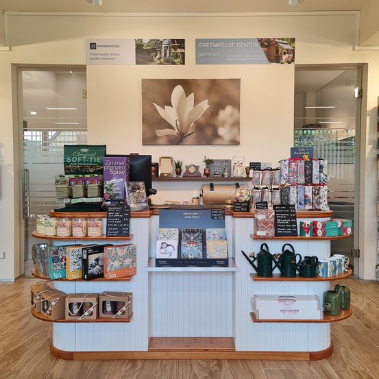 Das Innere eines Gewächshauses mit Holzregalen, gefüllt mit verschiedenen Produkten wie Büchern, Gießkannen und anderen Artikeln. Banner und Poster hängen an der Wand, und ein Bild einer Blume ist ausgestellt.