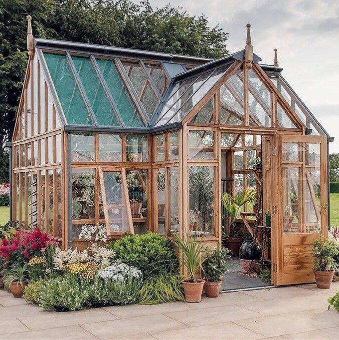 Ein Holzgewächshaus mit grünem Dach steht in einem Garten. Im Inneren sind verschiedene Topfpflanzen ausgestellt, und das Gewächshaus verfügt über Glasfenster und Türen.