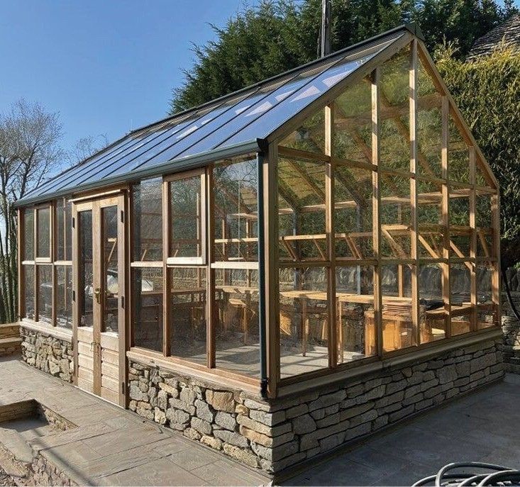 Ein Gewächshaus mit Glasdach und Holzrahmen, umgeben von einer Steinmauer, mit Holzbänken und Tischen im Inneren.