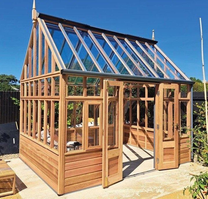 Ein Holzgewächshaus mit Glasdach und mehreren Fenstern steht im Freien. Die Struktur ist von einem Holzzaun umgeben.