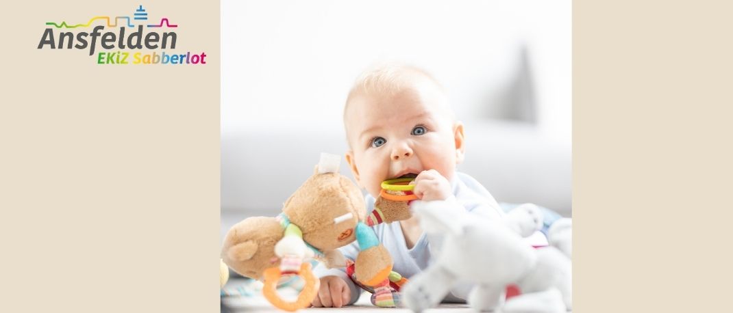 Ein Baby sitzt auf einem Bett und kaut auf einem Spielzeug, während es ein anderes Spielzeug in der Hand hält. Das Bild hat einen Wasserzeichen mit dem Text 'Ansfeldern'.
