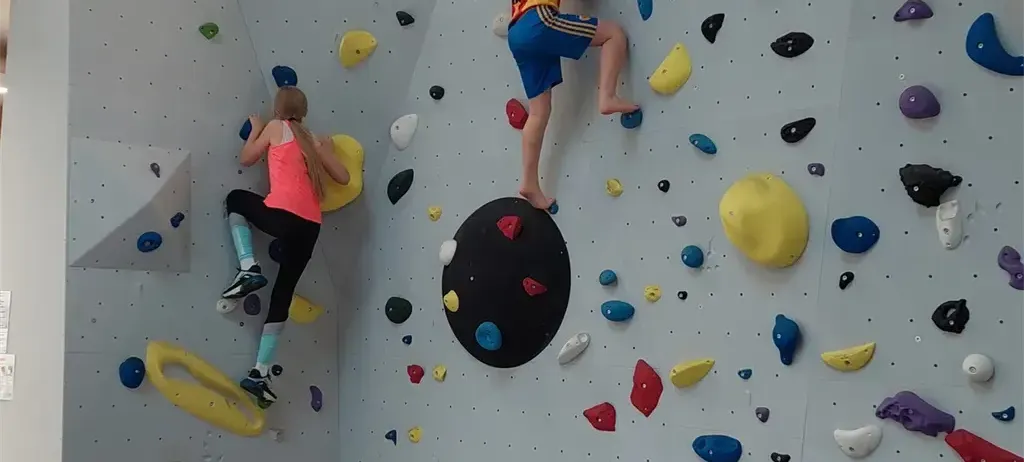 Zwei Personen klettern an einer Indoor-Kletterwand mit verschiedenen farbigen Griffen. Ein Kletterer steigt auf, während der andere mit einer gelben Schlaufe absteigt.