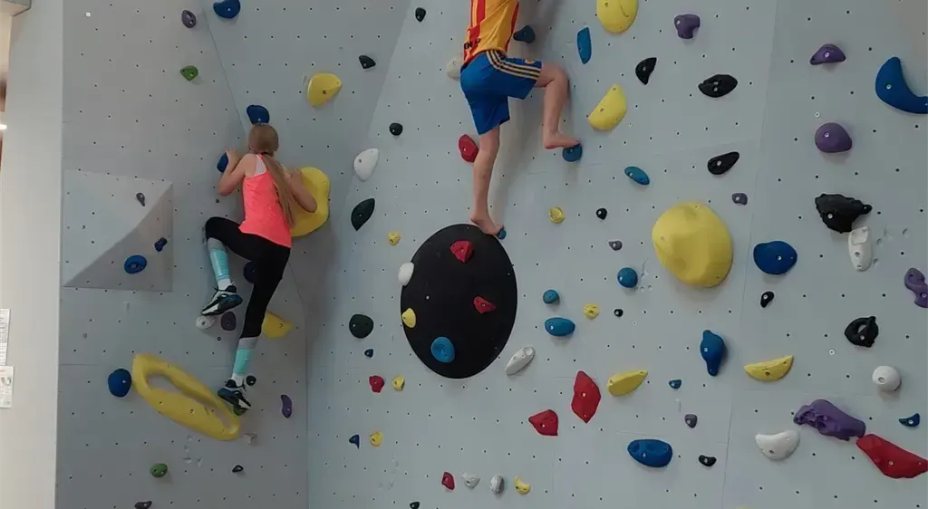 Zwei Personen klettern an einer Indoor-Kletterwand mit verschiedenen farbigen Griffen. Ein Kletterer steigt auf, während der andere mit einer gelben Schlaufe absteigt.