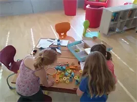 Drei Kinder spielen mit Spielzeug auf einem Holztisch in einem Spielzimmer mit mehreren Stühlen, einem Bücherregal und einem roten Boden. Das Mädchen in Pink sitzt, während die anderen stehen.
