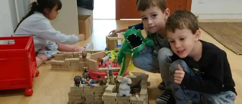 Drei Kinder spielen mit verschiedenen Tierspielzeug auf einem Holzboden. Ein Junge hält einen grünen Spielzeugkrokodil und ein anderer Junge zeigt Daumen hoch. Ein Mädchen sitzt neben ihnen. Neben dem Mädchen steht eine rote Spielzeugaufbewahrung.