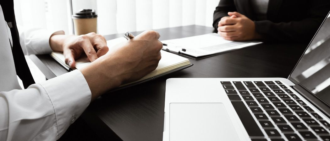 Eine Person sitzt an einem Schreibtisch mit Laptop und schreibt in ein Notizbuch. Gegenüber sitzt eine andere Person. Ein Kaffeebecher steht auf dem Schreibtisch.