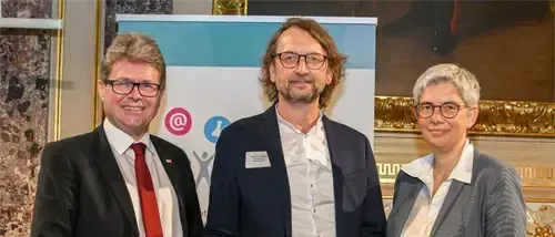 Drei Menschen in formeller Kleidung stehen lächelnd zusammen. Der Mann in der Mitte hält ein Zertifikat. Hinter ihnen ist ein Banner mit einem Logo und einem Symbol. Sie stehen vor einer Wand mit einem Bilderrahmen.
