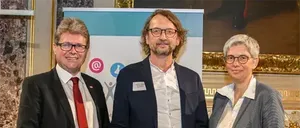 Drei Menschen in formeller Kleidung stehen lächelnd zusammen. Der Mann in der Mitte hält ein Zertifikat. Hinter ihnen ist ein Banner mit einem Logo und einem Symbol. Sie stehen vor einer Wand mit einem Bilderrahmen.