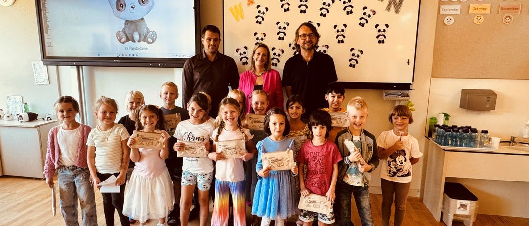 Eine Gruppe von Kindern und zwei Erwachsenen posiert für ein Foto in einem Raum mit einer Tafel dahinter. Die Kinder halten Urkunden in den Händen.