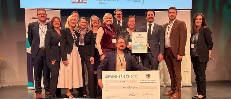 Mehrere Personen stehen auf einer Bühne mit einem Banner hinter ihnen, das 'Innovative Schulen Osterreichs' besagt. Ein Mann in der Mitte hält ein Zertifikat und einen Scheck.