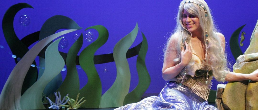 Eine Frau, die als Meerjungfrau verkleidet ist, sitzt auf der Bühne und trägt einen glänzenden blauen Schwanz und eine weiße Corsage. Sie lächelt und sieht in die Kamera. Der Hintergrund ist lila mit grünen Pflanzen.