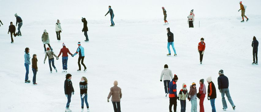 Eine Gruppe von Menschen eisskatet auf einem gefrorenen See, einige halten sich an den Händen und andere stehen allein. Sie sind in Winterkleidung gekleidet.