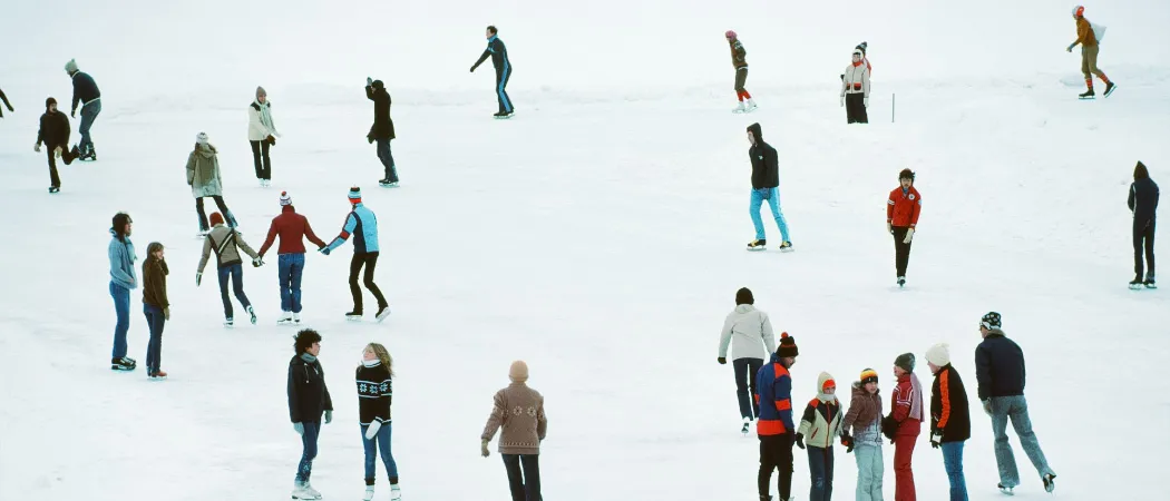Eine Gruppe von Menschen eisskatet auf einem gefrorenen See, einige halten sich an den Händen und andere stehen allein. Sie sind in Winterkleidung gekleidet.