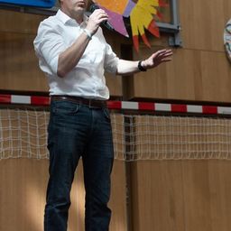 Ein Mann in einem weißen Hemd und Jeans hält eine Rede und hält ein Mikrofon in der Hand. Er steht auf einer Bühne mit einer hölzernen Wand und einem Netz dahinter.