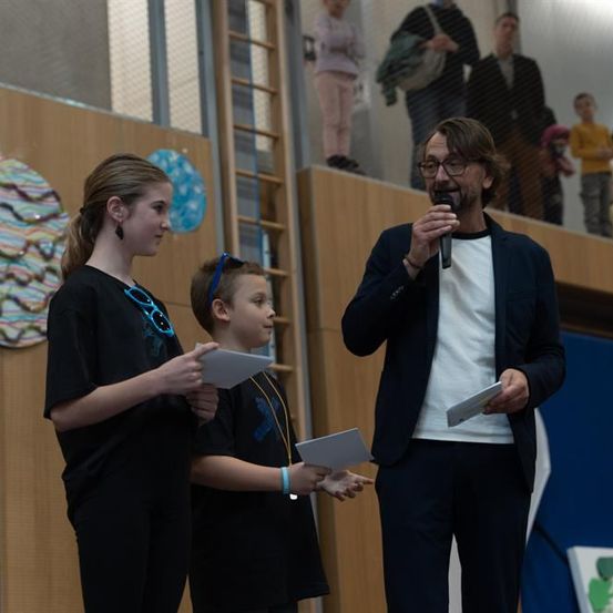 Ein Mann hält eine Rede und hält ein Mikrofon in der Hand. Zwei Kinder stehen neben ihm und halten Papiere. Dahinter stehen Leute auf einem Balkon.