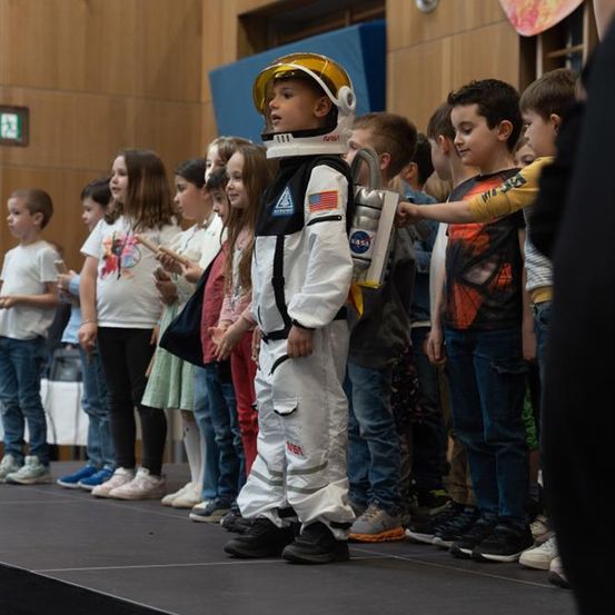 Eine Gruppe von Kindern, einige in Freizeitkleidung und andere in Kostümen, stehen auf einer Bühne, darunter ein Junge in einem NASA-Astronautenanzug.