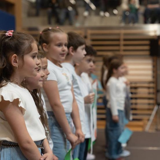 Mehrere kleine Kinder stehen in einer Reihe auf der Bühne, tragen weiße Hemden und halten grüne Bänder. Im Hintergrund sind einige weitere Kinder und Erwachsene zu sehen, die möglicherweise zuschauen.