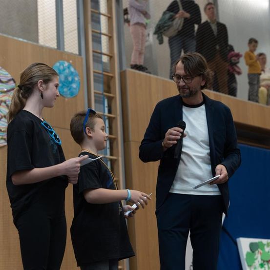 Ein Mann spricht in ein Mikrofon auf einer Bühne mit zwei Kindern, die Spielcontroller halten. Sie stehen vor einer Holzwand mit farbenfrohen Kunstwerken. Zuschauer stehen im Hintergrund.