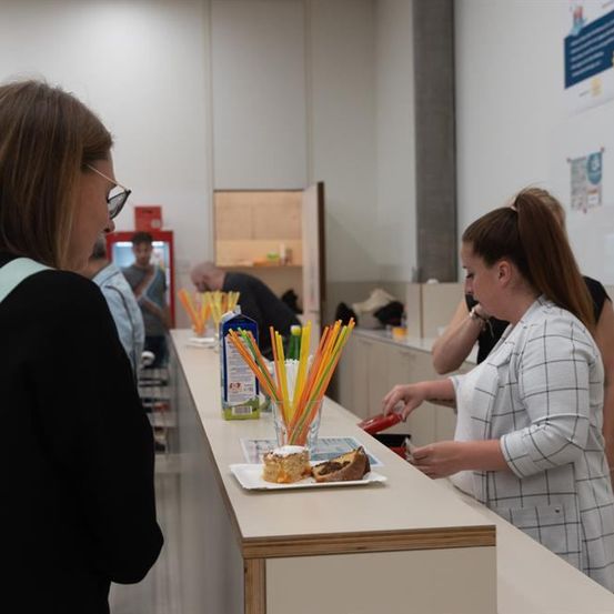 Eine Frau bereitet in einer Küche Essen zu, während eine andere Frau mit Brille zuschaut. Es gibt Desserts, Getränke und Utensilien auf dem Tresen.
