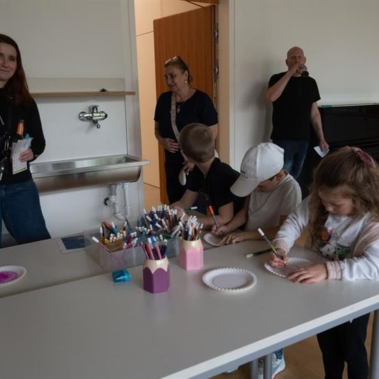 Drei Kinder und eine erwachsene Frau sitzen an einem Tisch und basteln. Die Frau hält ein Papier und schaut die Kinder an. Die Kinder zeichnen mit Stiften und Bleistiften auf Tellern.