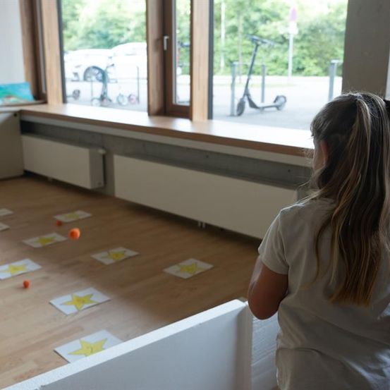 Ein junges Mädchen in einem weißen Shirt steht drinnen und blickt aus einem Fenster mit Blick auf einen Roller und Autos draußen. Drinnen sind gelbe Sterne auf dem Boden angeordnet.