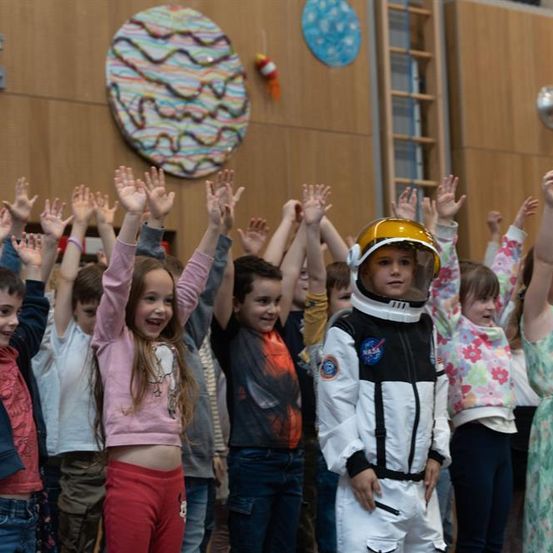 Eine Gruppe von Kindern in lässiger Kleidung und ein Kind in einem NASA-Astronautenanzug heben die Hände in einem Innenraum.