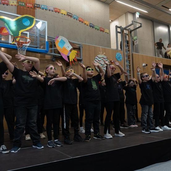 Eine Gruppe von Schülern in schwarzen Shirts und Hosen tritt auf einer Bühne auf, einige mit Brillen. Ein Basketballkorb und bunte Dekorationen sind dahinter.