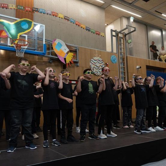 Eine Gruppe von Kindern in gleichen schwarzen T-Shirts steht auf einer Bühne in einer Turnhalle. Sie tragen Gläser mit bunten Linsen und machen Handgesten. Die Wände sind mit verschiedenen bunten Plakaten dekoriert.