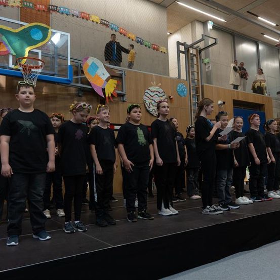 Eine Gruppe von Kindern steht auf der Bühne, einige halten Papiere in den Händen, tragen schwarze T-Shirts, mit Weltraum-Dekorationen im Hintergrund.