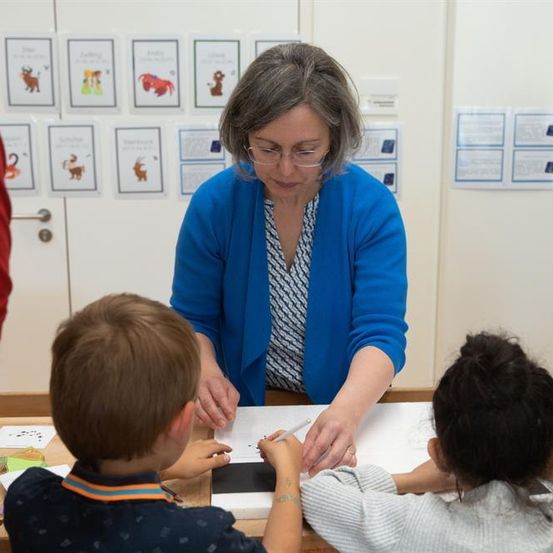 Eine ältere Frau hilft zwei Kindern bei ihren Hausaufgaben an einem Tisch im Klassenzimmer.