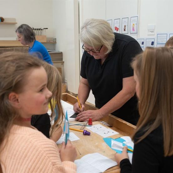 Eine ältere Frau mit Brille arbeitet an einem Bastelprojekt an einem Holztisch mit einer Gruppe von Kindern.