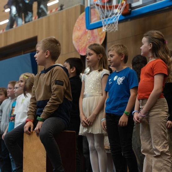 Mehrere Kinder stehen in einer Reihe in einer Turnhalle. Ein Junge sitzt auf einer Holzkiste. Ein Basketballkorb ist im Hintergrund.