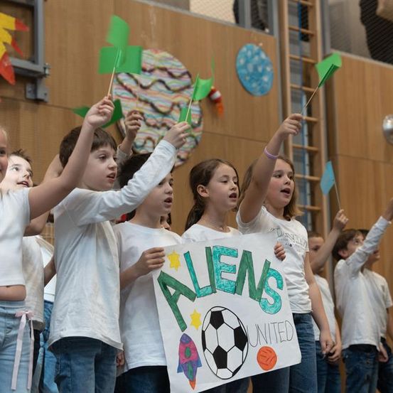 Eine Gruppe von Kindern in weißen T-Shirts hält ein Schild mit der Aufschrift Aliens United hoch und heben ihre Hände und Flaggen. Sie scheinen in einer Schulaula aufzutreten.