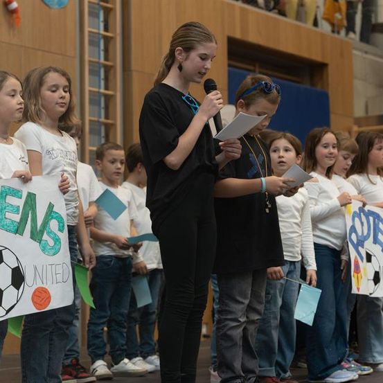 Eine Gruppe von Kindern steht in einer Reihe, einige halten Schilder und Fahnen. Ein Kind hält ein Mikrofon und Papier, und ein anderes hält ein Fußballschild. Sie scheinen bei einer Veranstaltung zu sein.