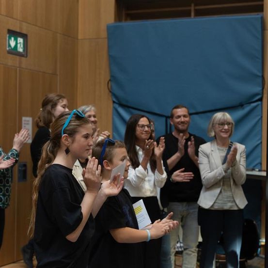 Mehrere Menschen stehen in einem Raum und klatschen. Eine Frau mit Brille hält ein Papier. Hinter ihnen ist eine blaue Wand teilweise mit einem Vorhang bedeckt.