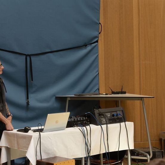 Ein Mann steht vor einem Schreibtisch mit einem Laptop, umgeben von verschiedenen elektronischen Geräten. Ein blauer Hintergrund und eine Holzwand sind im Hintergrund.