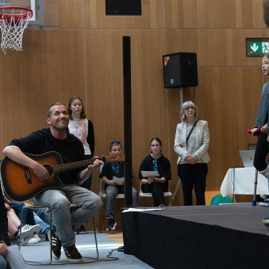 Ein Mann spielt Gitarre auf einer Bühne, während eine Gruppe von Menschen hinter ihm sitzt und steht, einige zuschauen und andere lesen. Ein Basketballkorb und Lautsprecher sind im Hintergrund.
