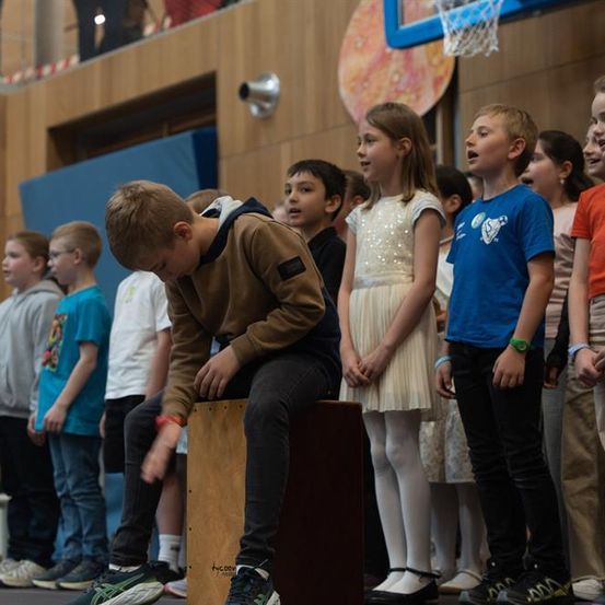 Eine Gruppe von Kindern steht in einem Raum, einige tragen Schuhe und andere nicht. Ein Junge spielt eine Trommel, während ein Mädchen in einem weißen Kleid hinter ihm steht.