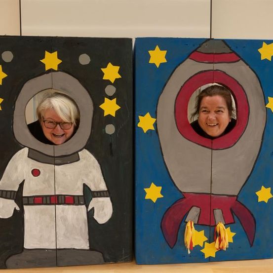 Zwei Frauen posieren durch Ausschnitte in bemalten Pappkartons. Eine ist eine Rakete, die andere ein Astronaut. Beide tragen verspielte Ausdrücke.