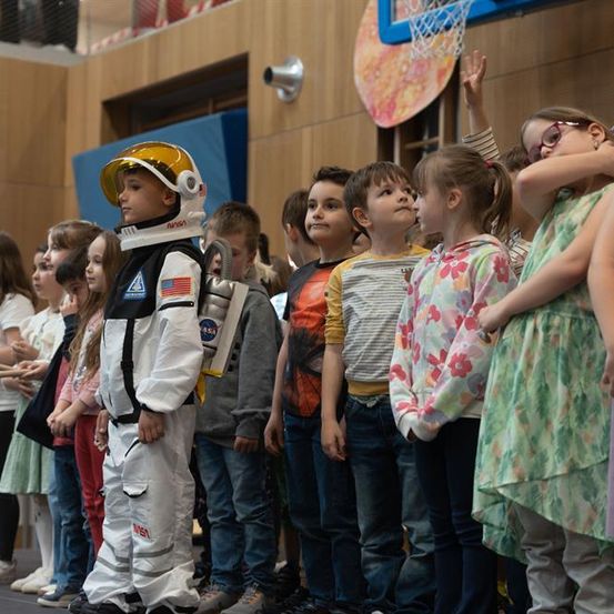 Ein junger Junge in einem NASA-Raumsuit steht inmitten einer Gruppe von Kindern in einem Raum, einige schauen mit Neugier nach oben.