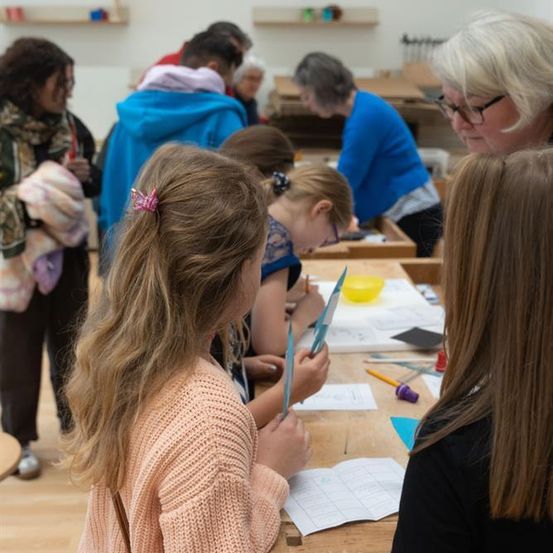 Eine Gruppe von Kindern und Erwachsenen versammelt sich um einen Tisch, wobei eine Frau mit Brille beobachtet. Sie sind in ein Bastelprojekt vertieft und verwenden Materialien wie Papier und Scheren. Die Kinder tragen Freizeitkleidung, und einige Erwachsene tragen Brillen.