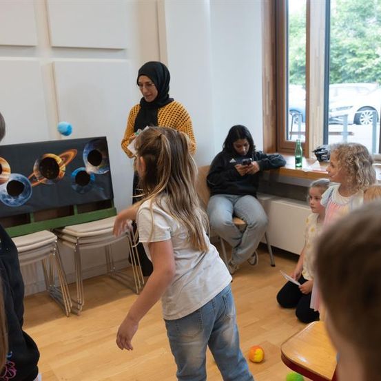Eine Gruppe von Kindern spielt ein Spiel in einem Raum mit Holzböden. Eine Frau steht vor einem Planetenposter, während ein Mädchen ihr Telefon betrachtet.