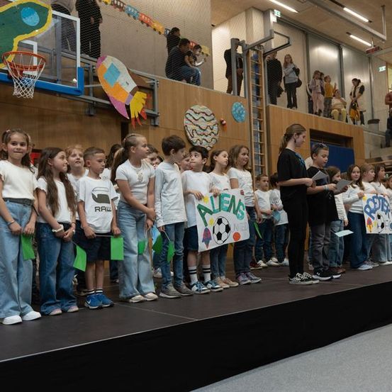 Kinder in weißen T-Shirts halten Schilder und stehen auf einer Bühne, während eine Frau in Schwarz zu ihnen spricht. Dahinter stehen andere auf einem Balkon, einige beobachten.