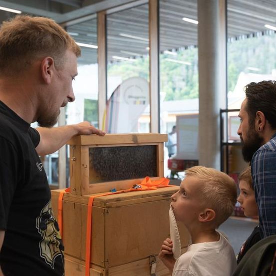 Ein Mann in einem schwarzen T-Shirt zeigt zwei Jungen und einem Mann in einem blauen Hemd eine Holzkiste. Die Kiste hat einen transparenten Deckel mit einem Bienenstock darin.