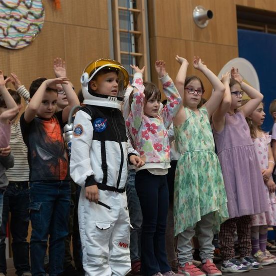 Eine Gruppe von Kindern steht auf der Bühne, eines in einem NASA-Astronautenanzug, während andere die Hände heben. Die Kinder sind in verschiedenen Outfits gekleidet, und die Umgebung hat Holzwände und einen blauen Hintergrund.