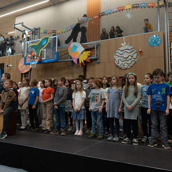 Eine Gruppe von Kindern steht auf einer Bühne in einer Turnhalle, einige stehen auf der linken Seite, ein Basketballkorb hinter ihnen und eine Menge auf der oberen Etage.