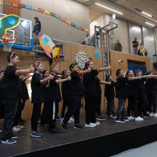 Eine Gruppe von Kindern in schwarzen Outfits führt auf einer Bühne auf, mit einem Basketballkorb im Hintergrund.