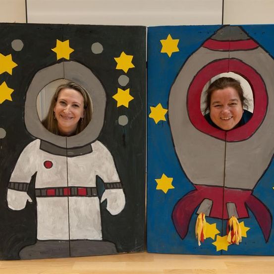 Zwei Frauen posieren in Ausschnitten, einer als Astronaut und der andere als Rakete, beide mit gelben Sternen. Die Astronauten-Schablone ist schwarz mit weißen und roten Details, während die Raketen-Schablone blau und grau mit roten und gelben Details ist.